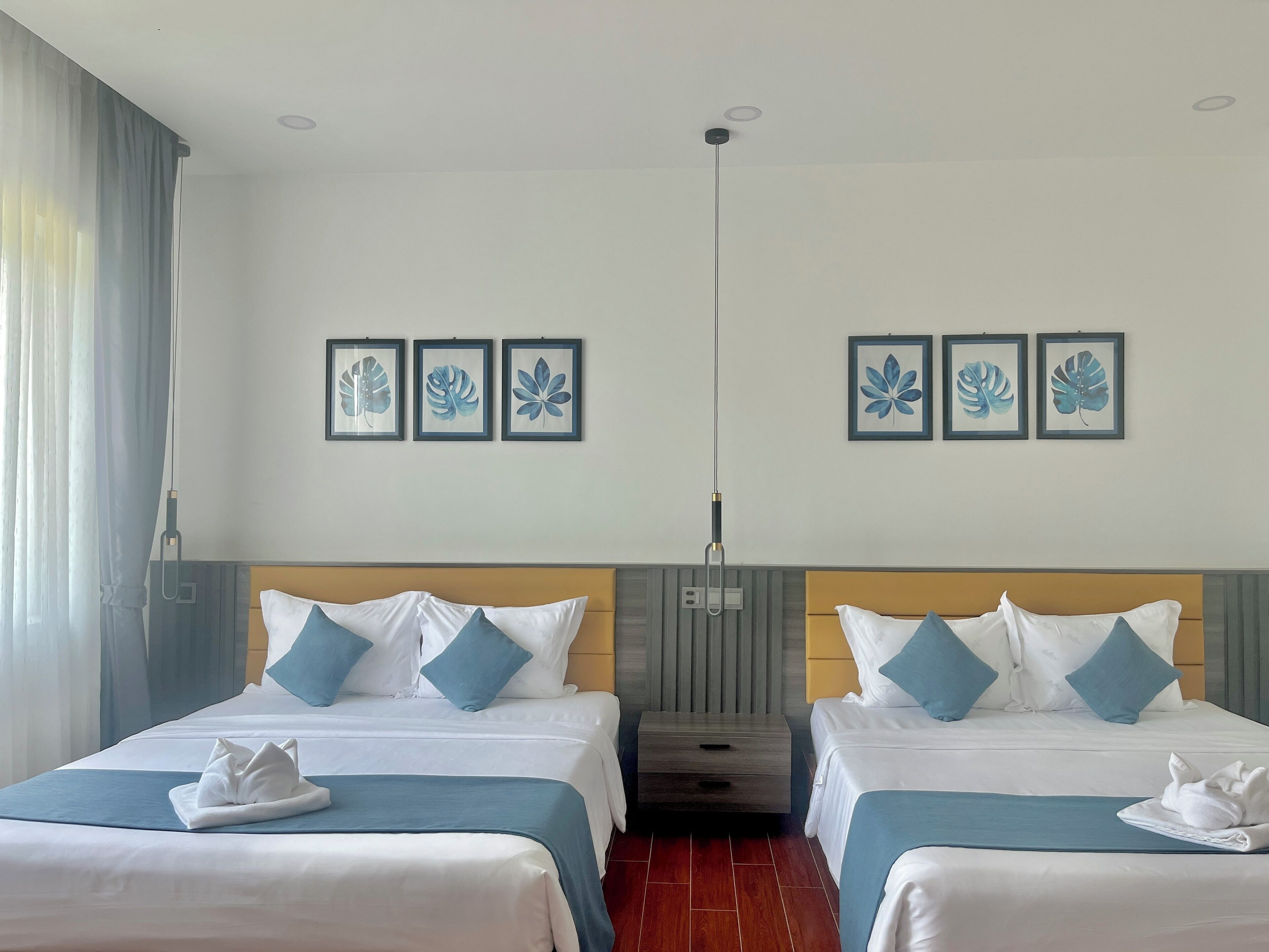 Habitación de hotel con dos camas, cada una con ropa blanca y almohadas azules, un cabecero de madera, una mesita de noche y una lámpara fijada a la pared.