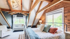 Une chambre avec un plafond en pente, un lit recouvert d’une literie à motifs, une table de chevet et une fenêtre donnant sur un paysage verdoyant.