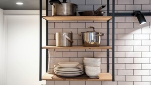 A kitchen with a black metal shelf unit holding pots and pans, a wooden shelf with plates, and a hanging light fixture. - TAKETO STAY KOTONI 102 (Sapporo)