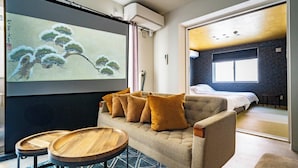 A modern hotel room with a sofa, two wooden side tables, a large screen displaying artwork, a bed, and a view of the outside. - TAKETO STAY KOTONI 102 (Sapporo)