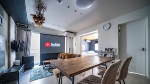 A modern living room with a wooden dining table, comfortable seating, a large screen displaying YouTube, and a view of the city. - TAKETO STAY KOTONI 102 (Sapporo)