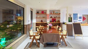 Uma área de refeições moderna com uma mesa de madeira comprida, cadeiras e um vaso central com flores. Estantes com objetos decorativos e um menu em lousa são visíveis.