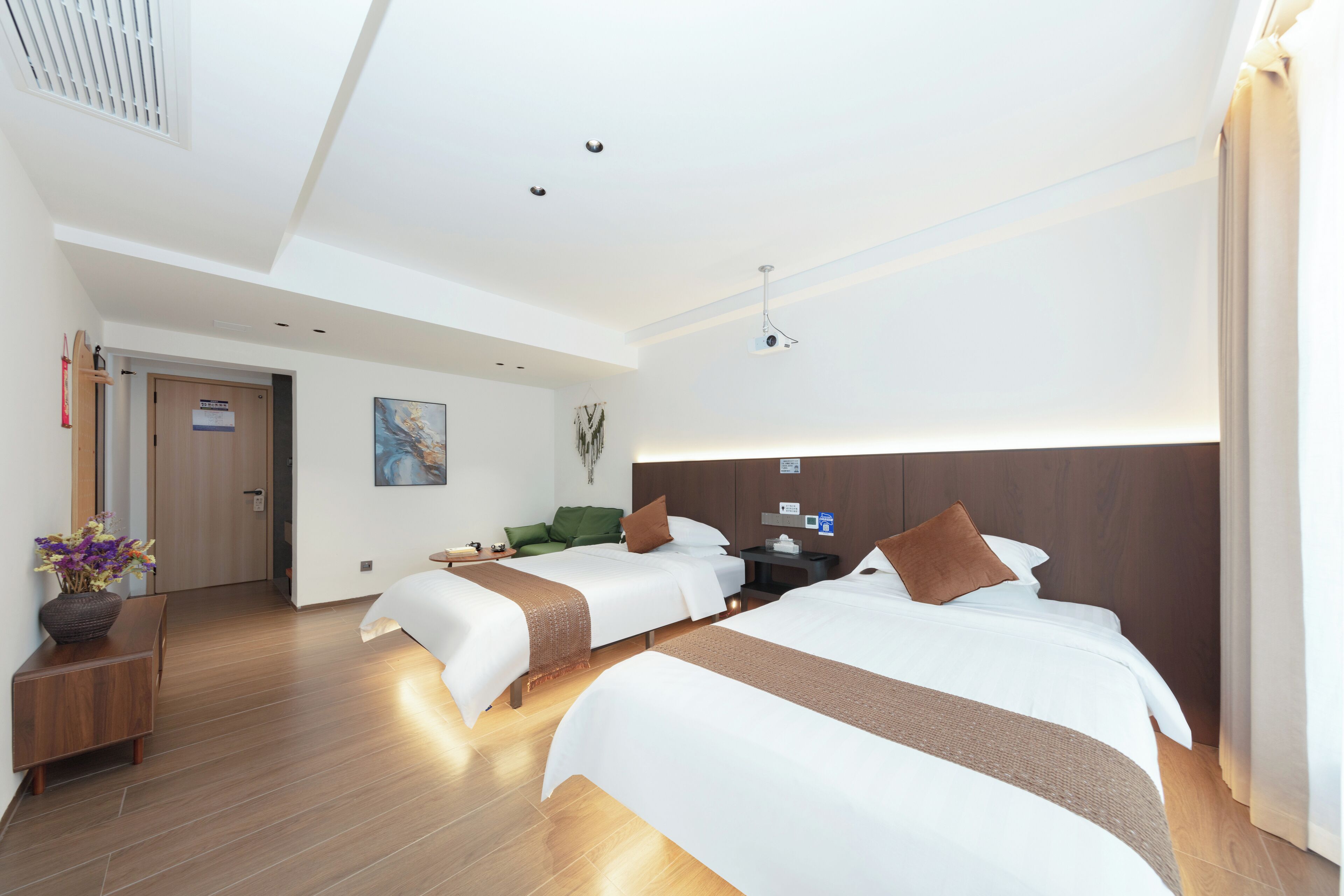 A hotel room with two beds, a wooden floor, a ceiling light, and a door.