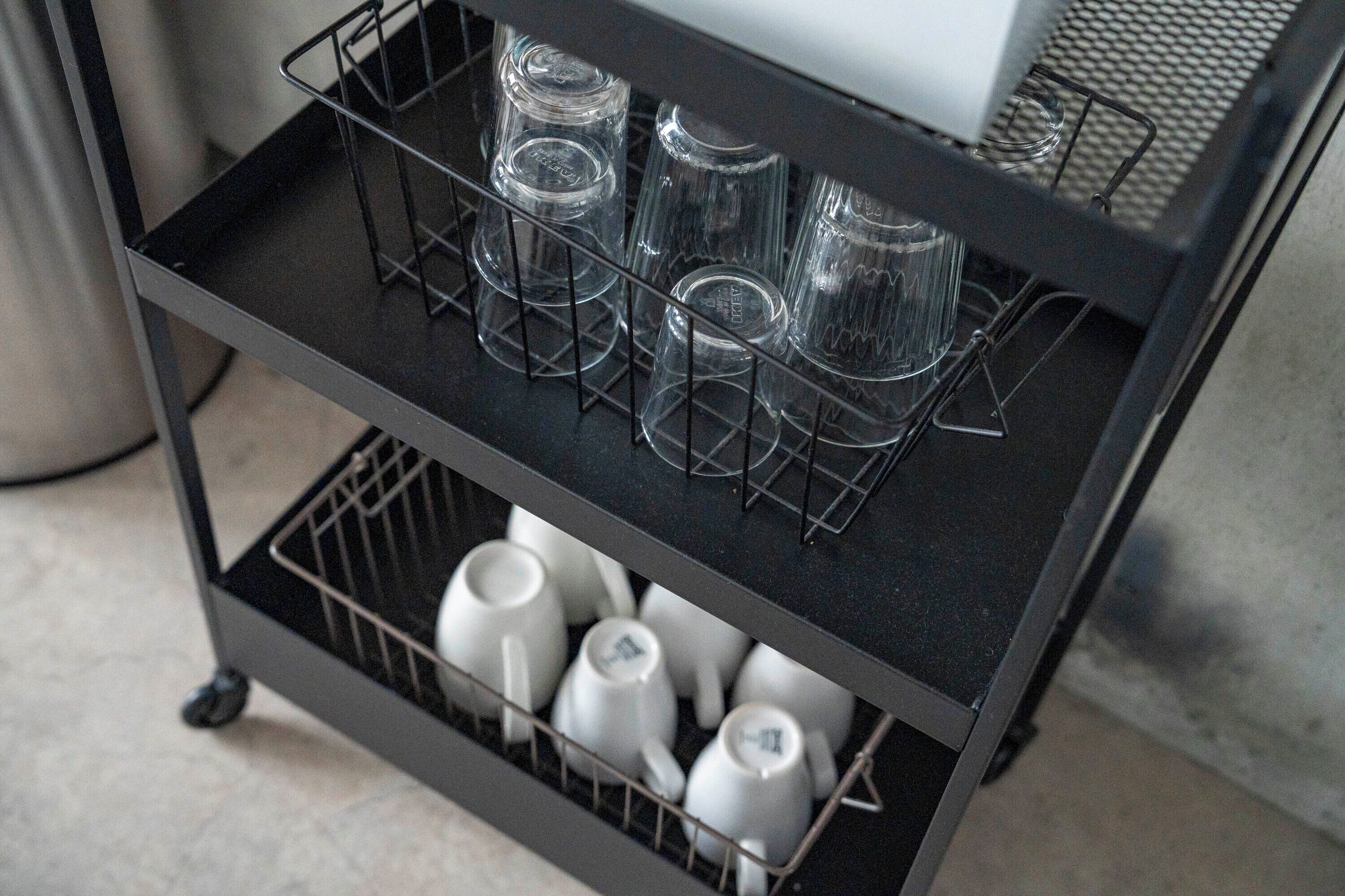 A black metal cart with two shelves, one holding clear glasses and the other holding white mugs.