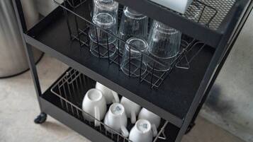 A black metal cart with two shelves, one holding clear glasses and the other holding white mugs.