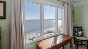 A room with a large window overlooking a beach, a wooden desk, and a chair with a cushion.