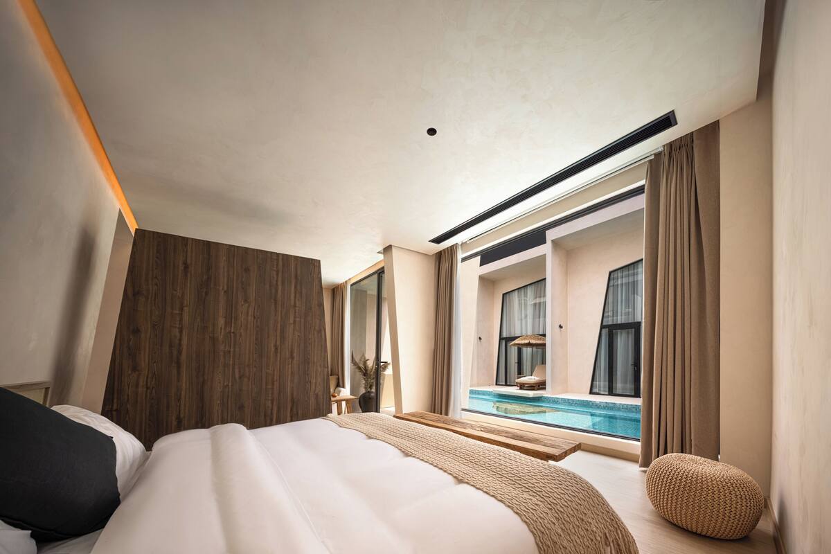 Hotelkamer met groot bed, houten hoofdeinde, uitzicht op een zwembad en een groot raam met gordijnen.