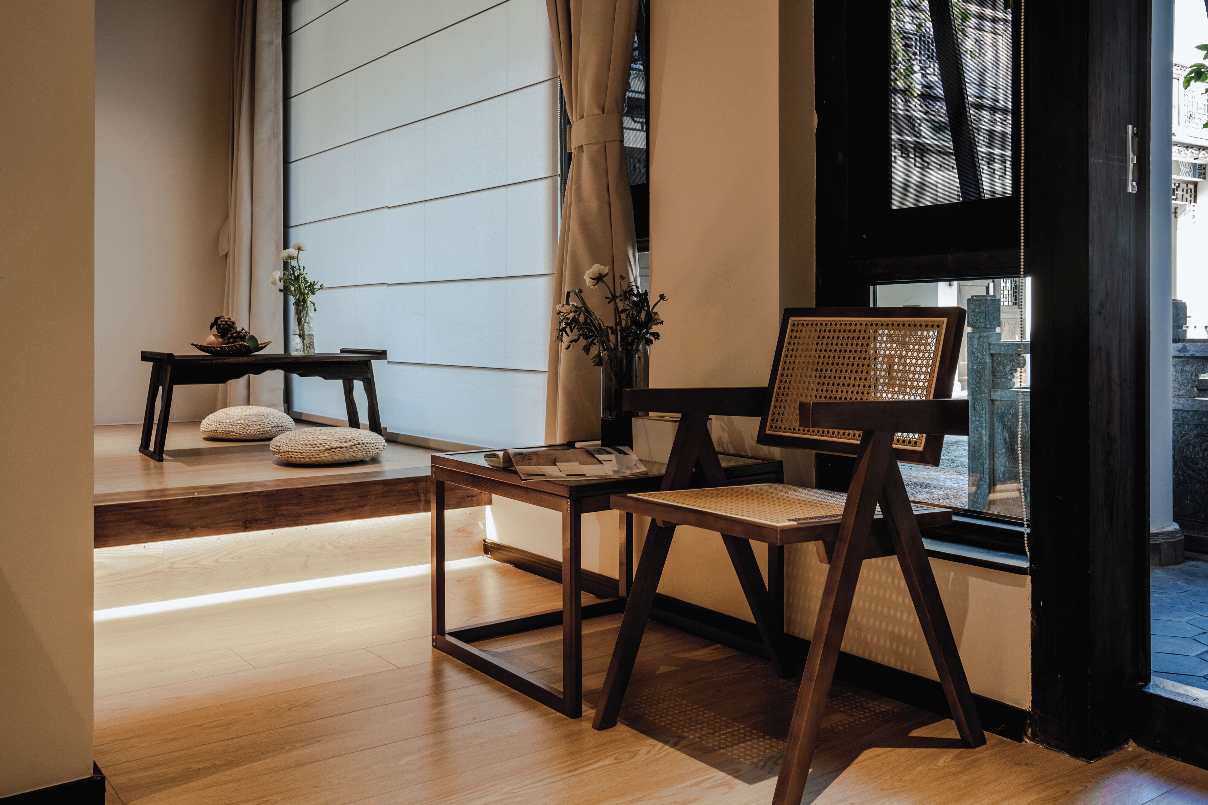 Una habitación moderna con piso de madera, una mesa de madera con un florero y una silla de mimbre, y vista a un edificio tradicional a través de una ventana.