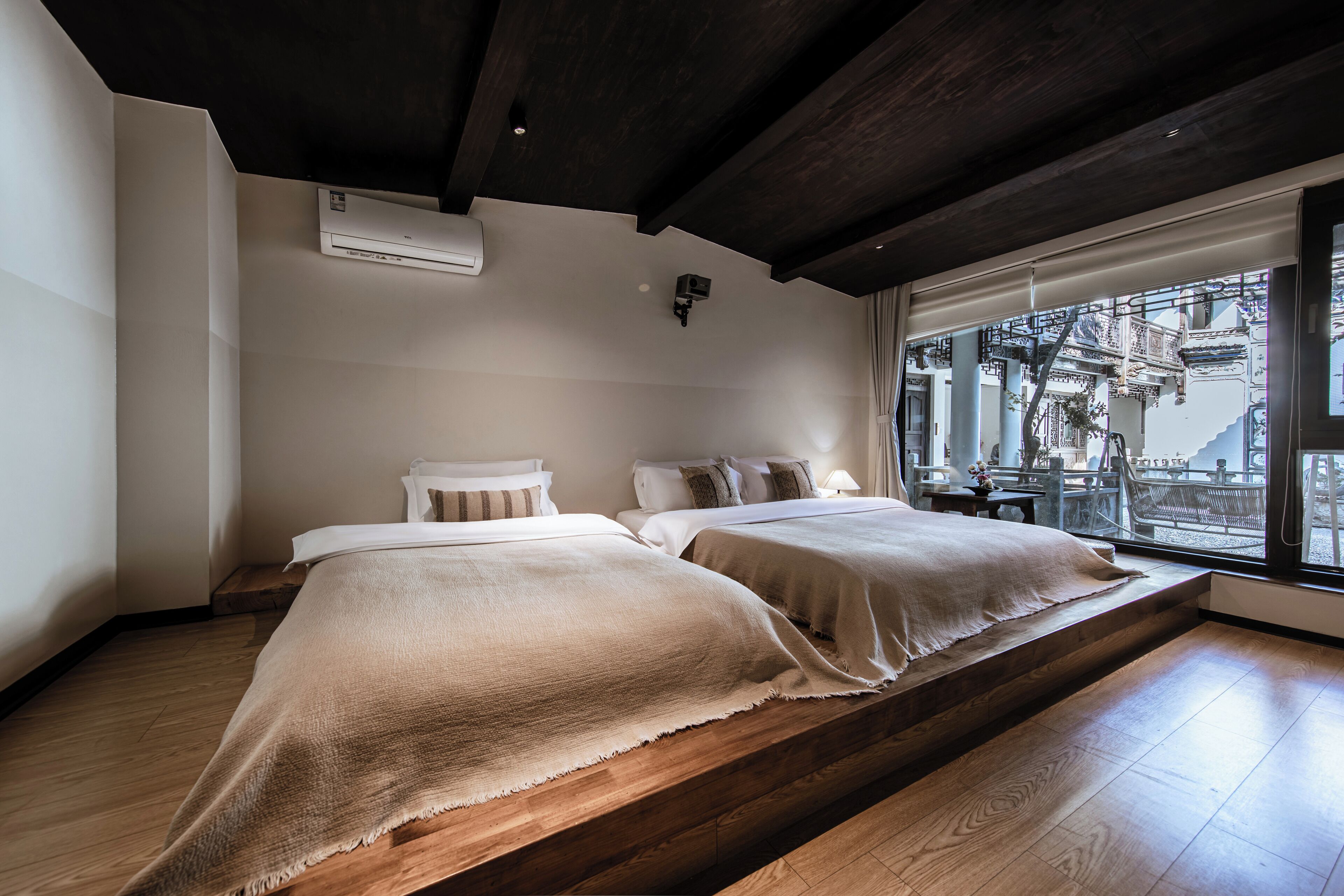 Un dormitorio con dos camas, suelo de madera, techo oscuro y una ventana con vistas a muebles de exterior.