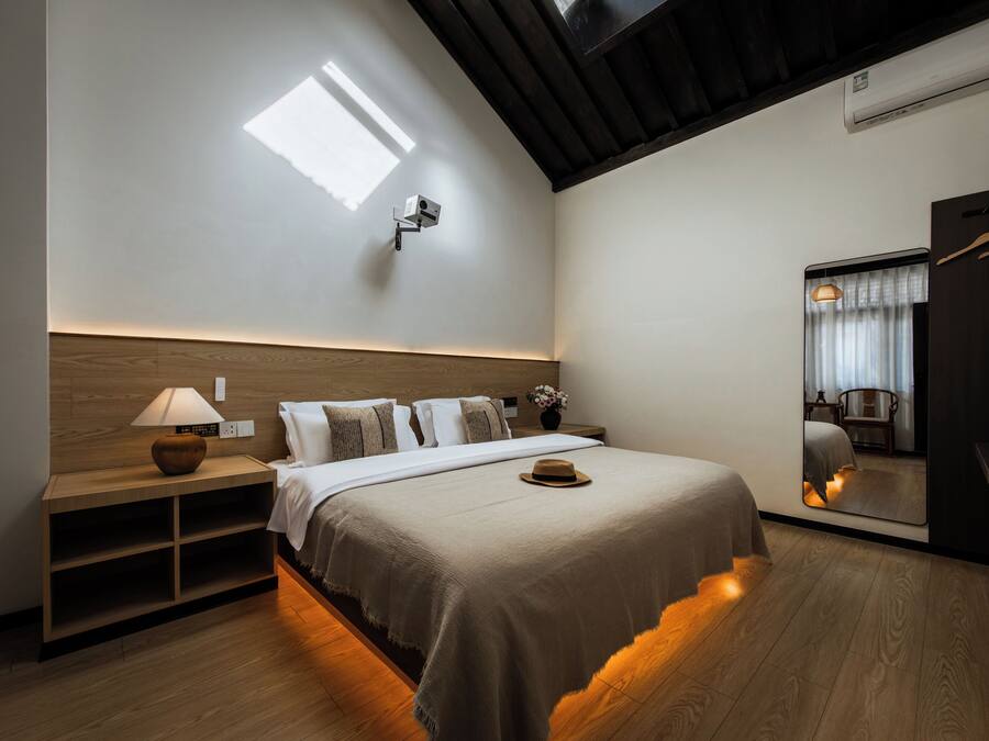 A modern bedroom with a wooden headboard, a bedside table with a lamp, a large bed with a beige blanket, a mirror, and a hanging coat rack.