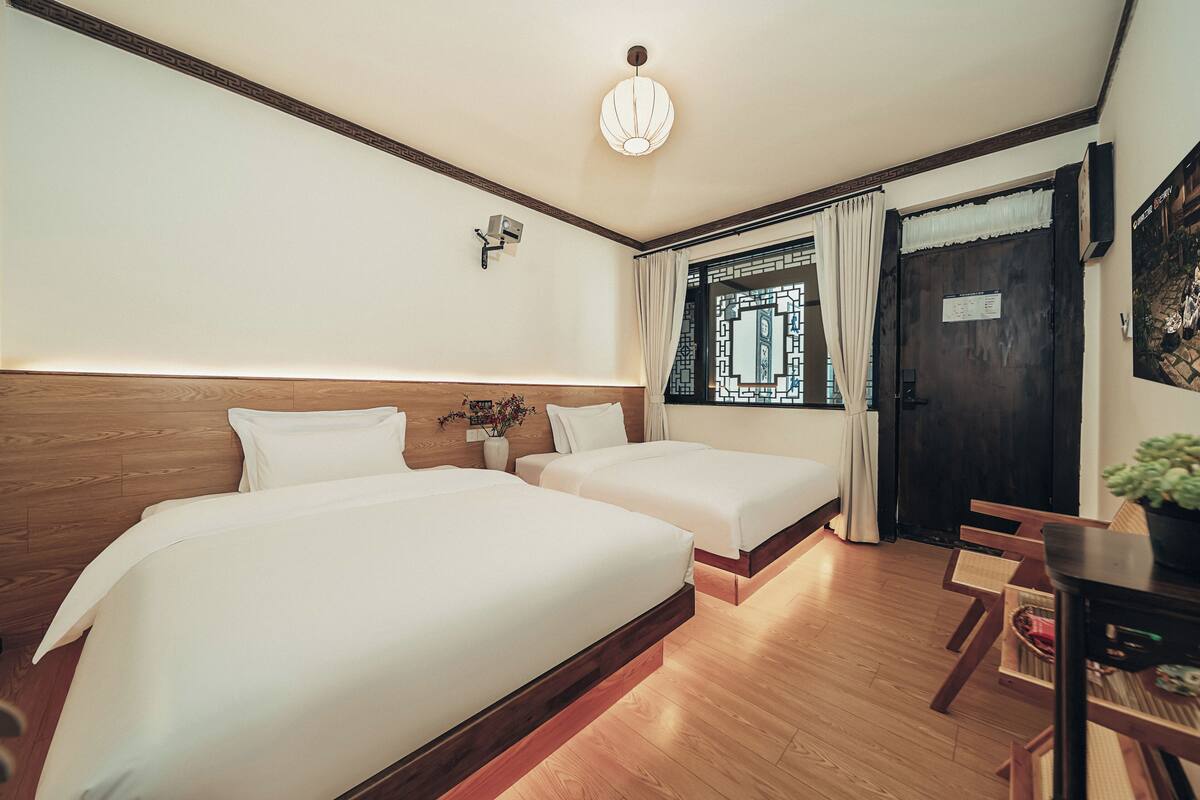 Habitación de hotel con dos camas, piso de madera, lámpara de techo, ventana con cortinas y puerta con un cartel.