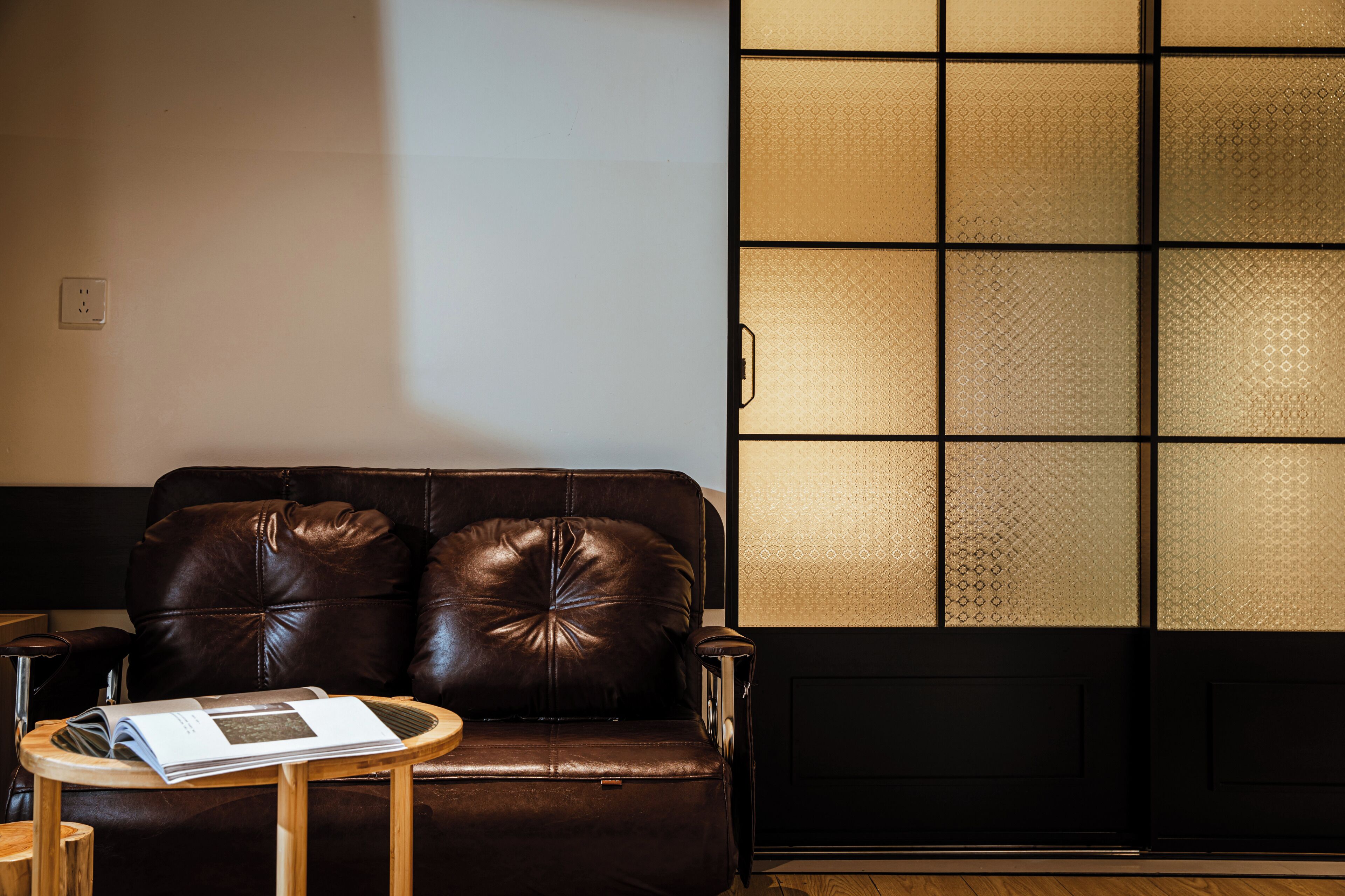 A brown leather sofa with a small wooden side table, a black door with a patterned glass panel, and a white wall with a power outlet.