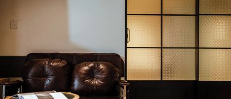 A brown leather sofa with a small wooden side table, a black door with a patterned glass panel, and a white wall with a power outlet.