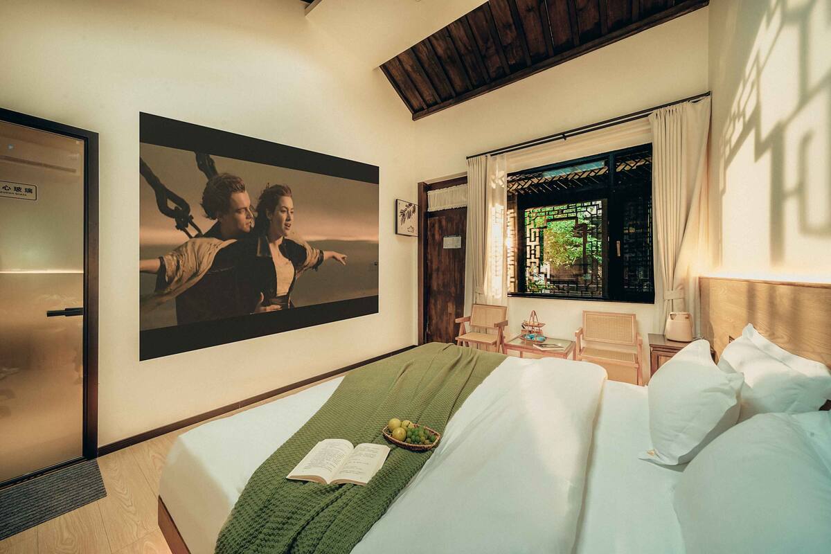 Una habitación de hotel con una cama, una manta verde, un libro abierto, una cesta de frutas y un cuadro grande en la pared.
