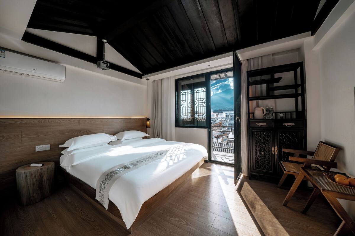 Un dormitorio con una cama grande, suelo de madera, techo con vigas a la vista, una mesita de noche, una silla, una pequeña mesa y vistas a través de una ventana.