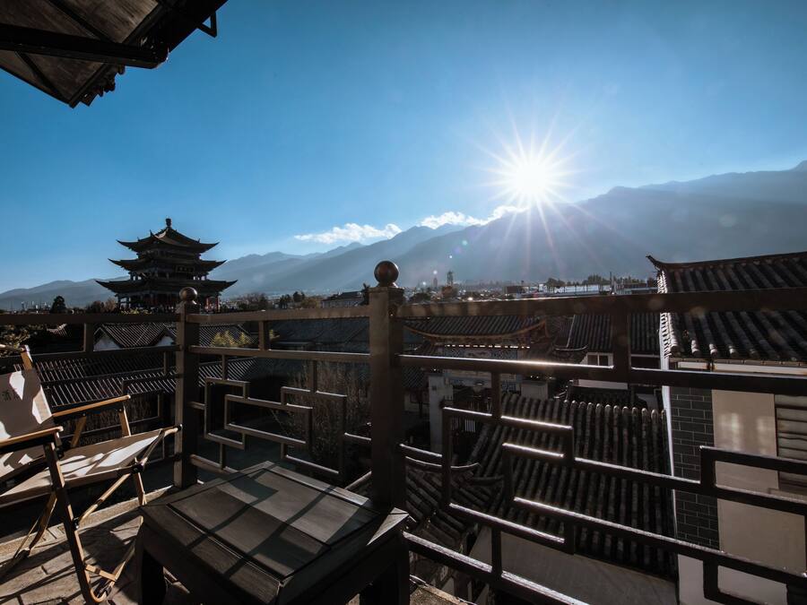 Un entorno arquitectónico tradicional chino con edificios de estilo pagoda escalonada, cielo despejado y una montaña iluminada por el sol al fondo.