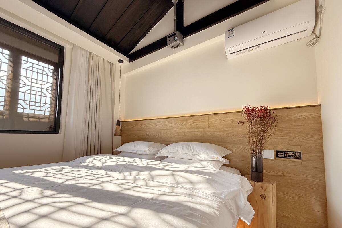 Una cama bien hecha con sábanas blancas, un cabecero de madera y una mesita de noche con un jarrón de flores secas.
