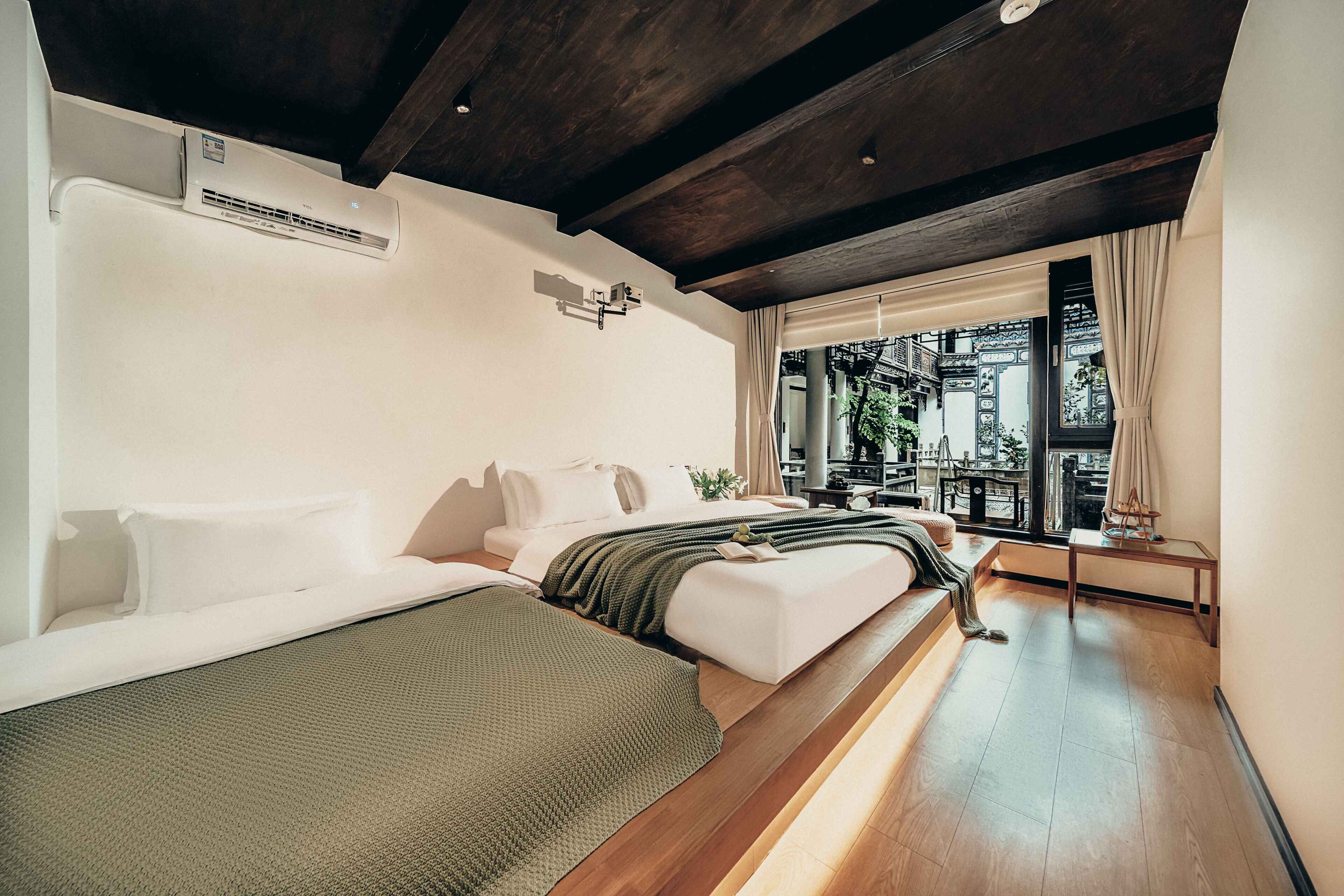 A modern hotel room with a wooden ceiling, a large window with curtains, a bed, a bedside table, and a small balcony.