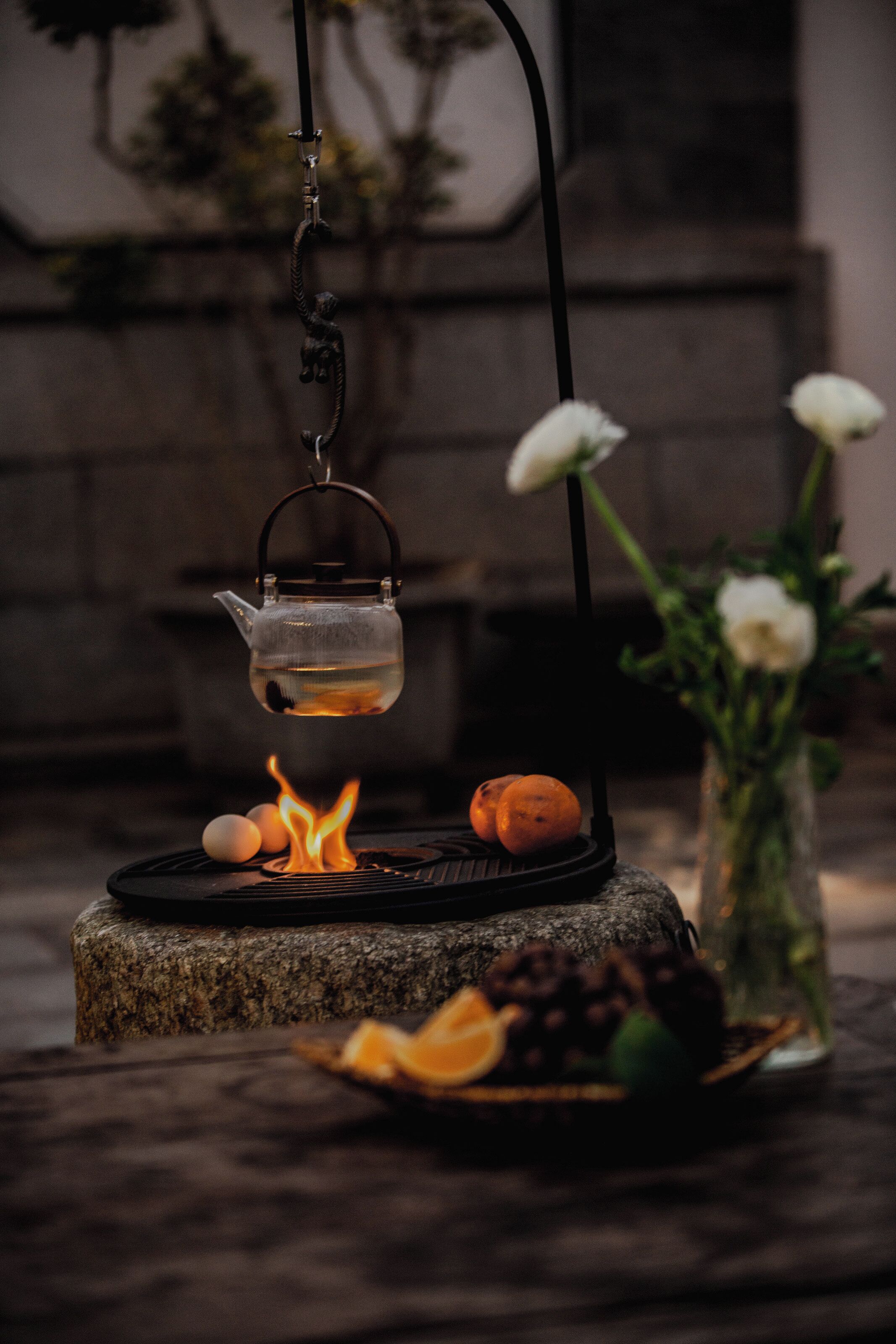 A tea kettle suspended over a charcoal grill with fruits and flowers on a stone table.