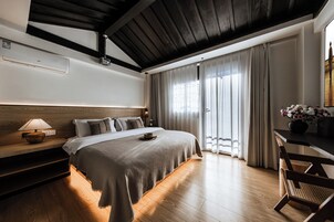 Un dormitorio con una cama grande, suelo de madera, techo con vigas a la vista, una mesita de noche con lámpara y vistas a un balcón con cortinas.