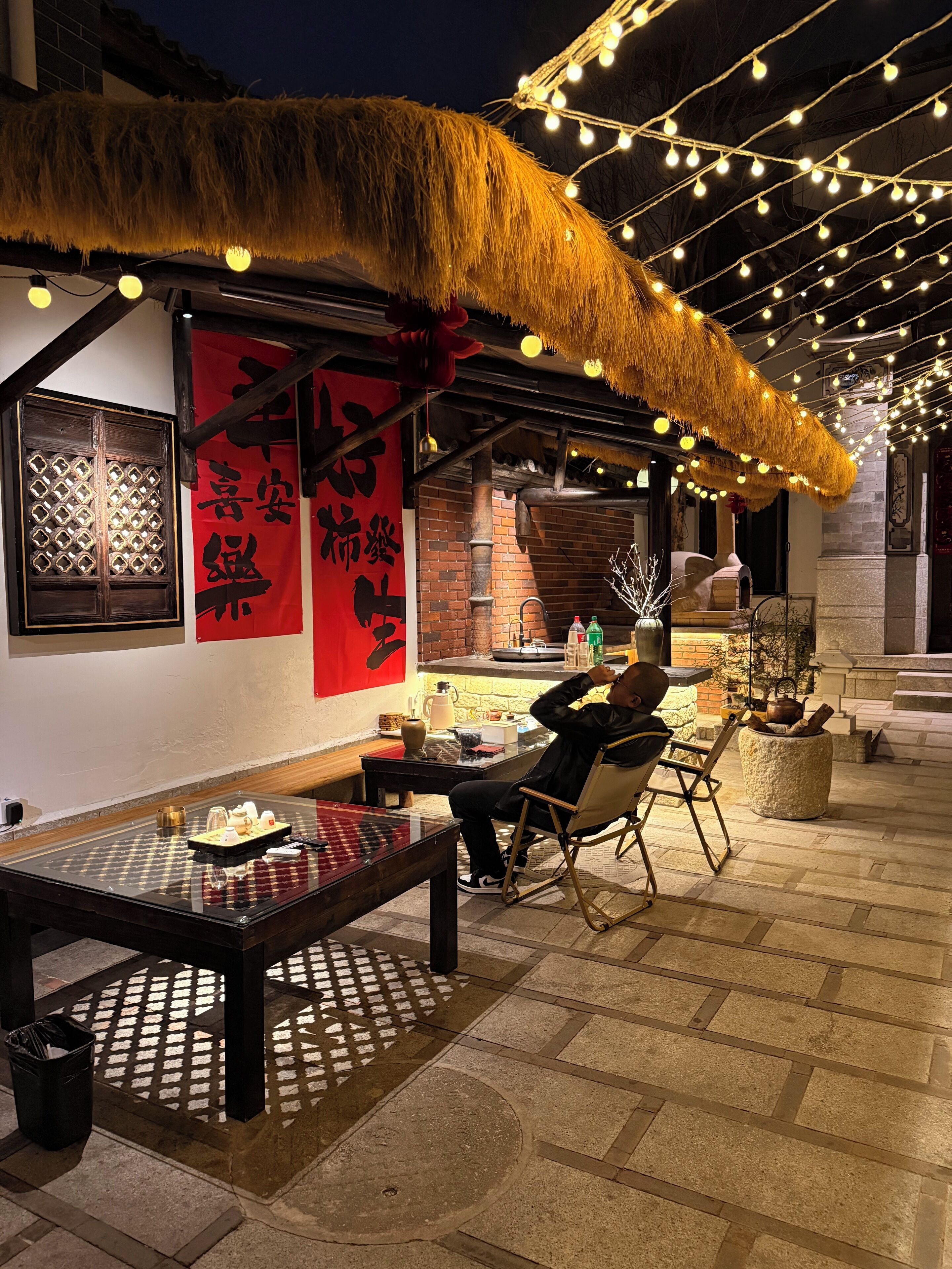 An outdoor dining area with a table and chairs, a thatched roof, string lights, and traditional decor.
