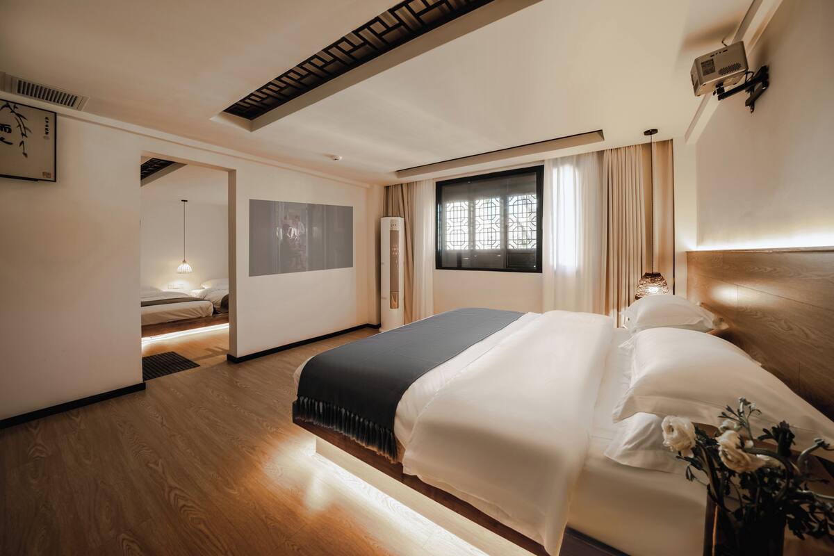 Una habitación de hotel moderna con una cama grande, una mesita de noche con lámpara, una ventana con cortinas y un techo con detalles de madera.