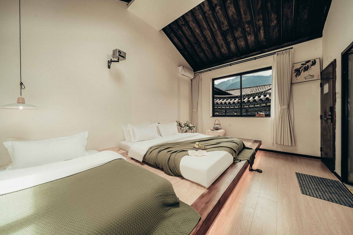 Habitación de hotel con techo de madera, un ventanal con vistas a la arquitectura tradicional, una cama con ropa blanca y una manta verde.