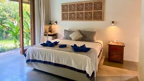 A bedroom with a large bed, a nightstand, a wall-mounted decorative panel, and a view of greenery through a sliding door. - Villa Kevin - 3BR Private Villa (Kecamatan Mengwi)
