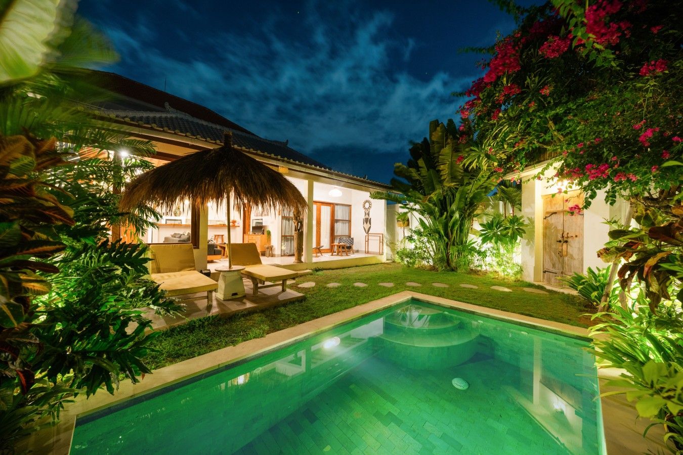A pool area with a thatched roof structure, outdoor seating, and lush greenery.