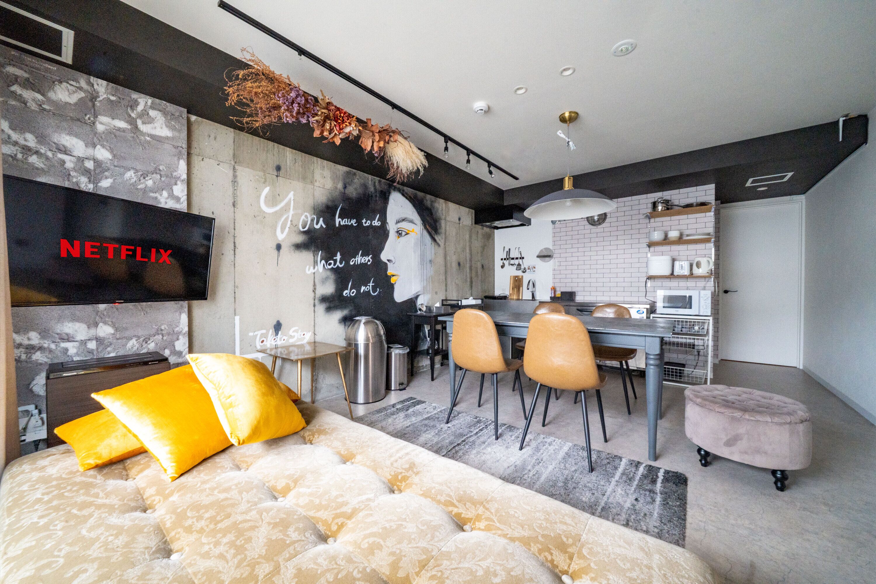 A modern living room with a large TV, a sofa with yellow cushions, a dining area with chairs, and a kitchen in the background.