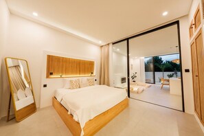 A modern bedroom with a large bed, a mirror, a wooden headboard, and a sliding glass door leading to an outdoor area. - Alcove Boutique Villa C 1BR (Kecamatan Kuta Selatan)