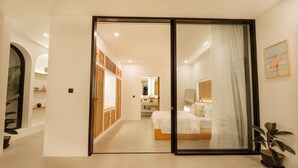 Habitación de hotel con puerta de vidrio corredera que da a un baño, una cama con ropa blanca, una mesita de noche y una planta en maceta.