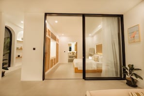 A hotel room with a sliding glass door leading to a bathroom, a bed with white bedding, a bedside table, and a potted plant.