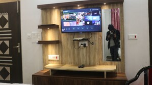 A modern living room with a flat-screen TV mounted on a wooden wall, a wooden bench, and a mirror reflecting a person taking a photo. - Narayan Marriage Hall And resort  (Sitamarhi)