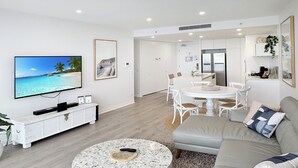 A modern living room with a large flat-screen TV, a round coffee table, a sofa, and a dining area with a white table and chairs. - Luxury Oracle Tower 2 Ocean Views 2BR Lvl33 (Broadbeach)