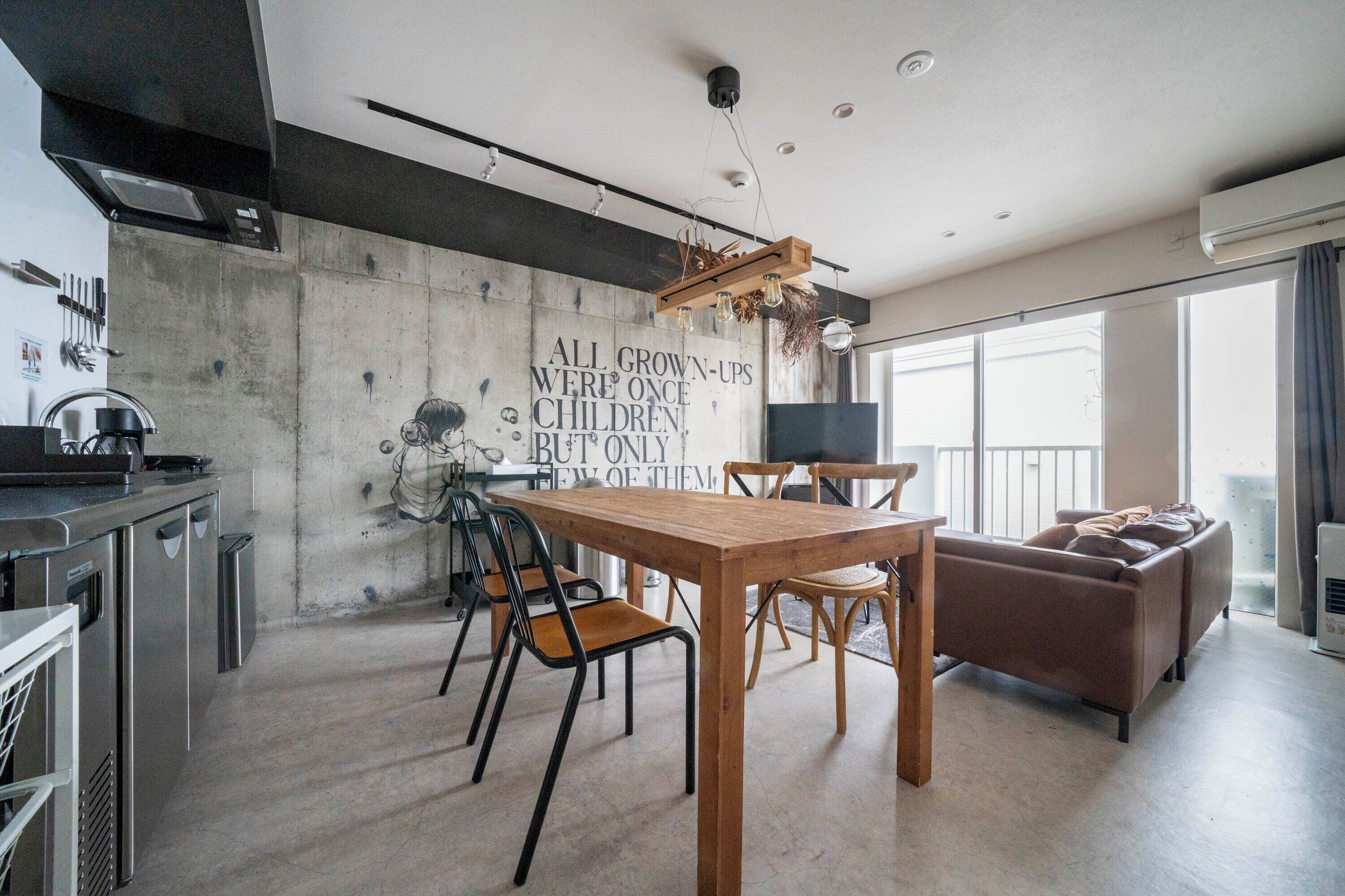 A modern kitchen with a concrete wall, wooden table, and chairs. There is a sofa and a TV in the living area.