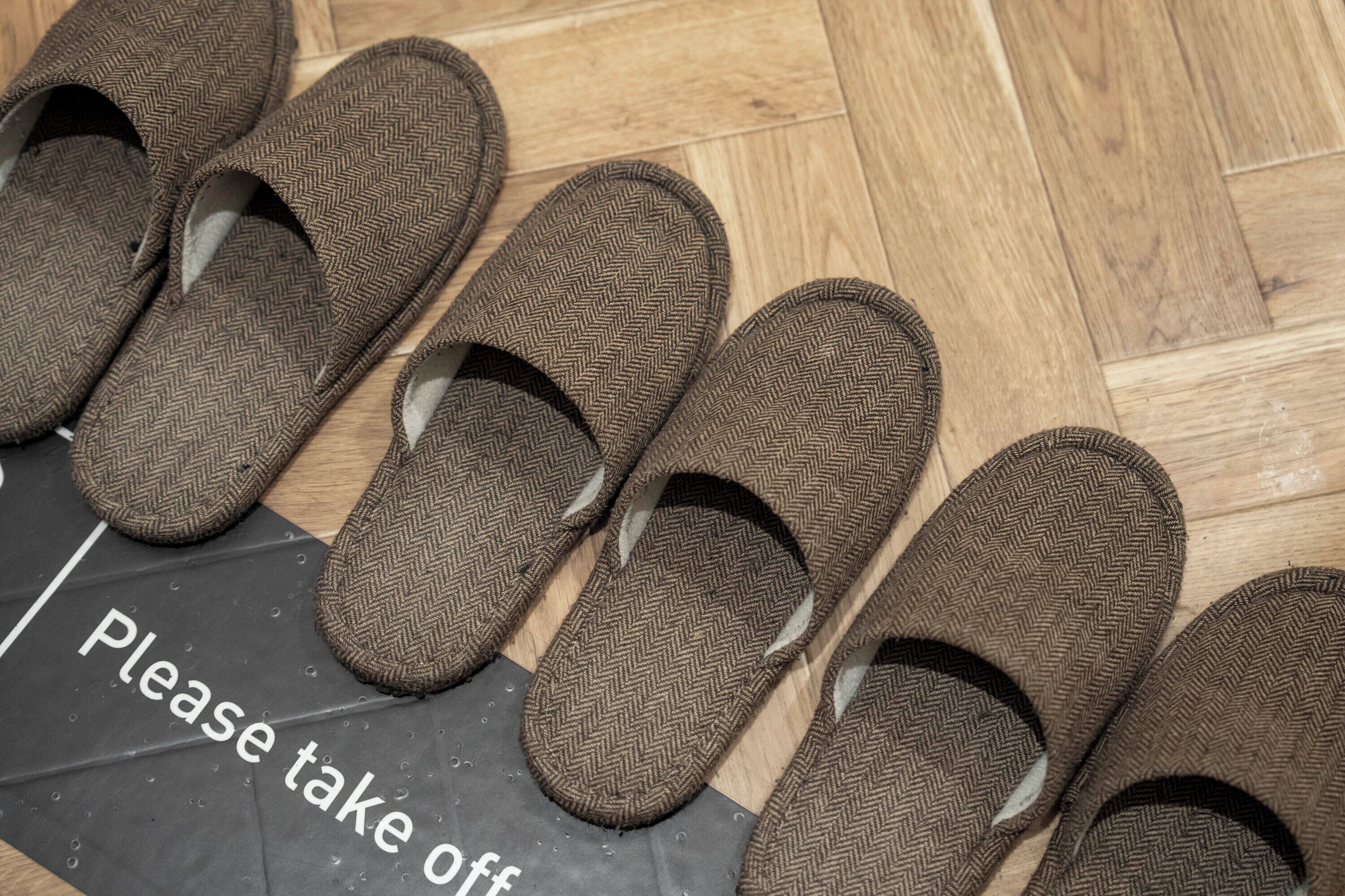 A pair of brown slippers with a herringbone pattern on a wooden floor next to a sign that says "Please take off."