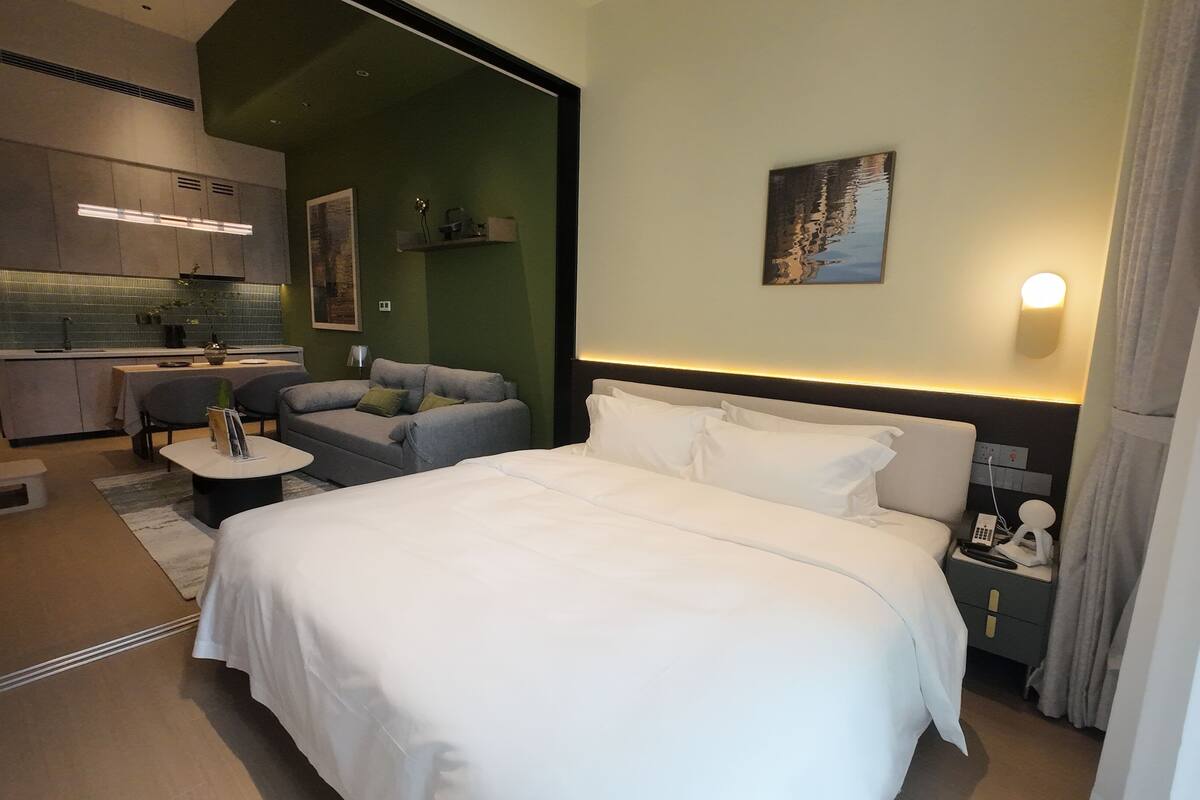 A hotel room with a large bed, a small table, a sofa, and a kitchenette in the background.