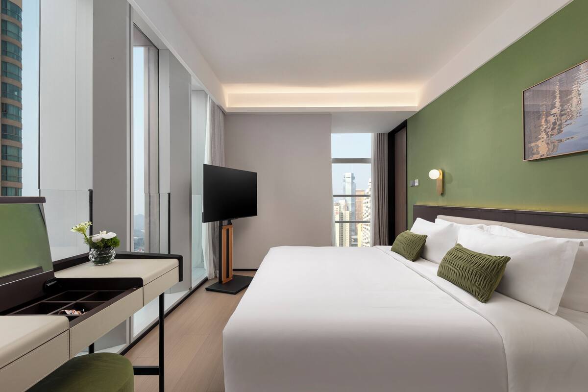 A hotel room with a bed, a desk, a television, and a window with a city view.