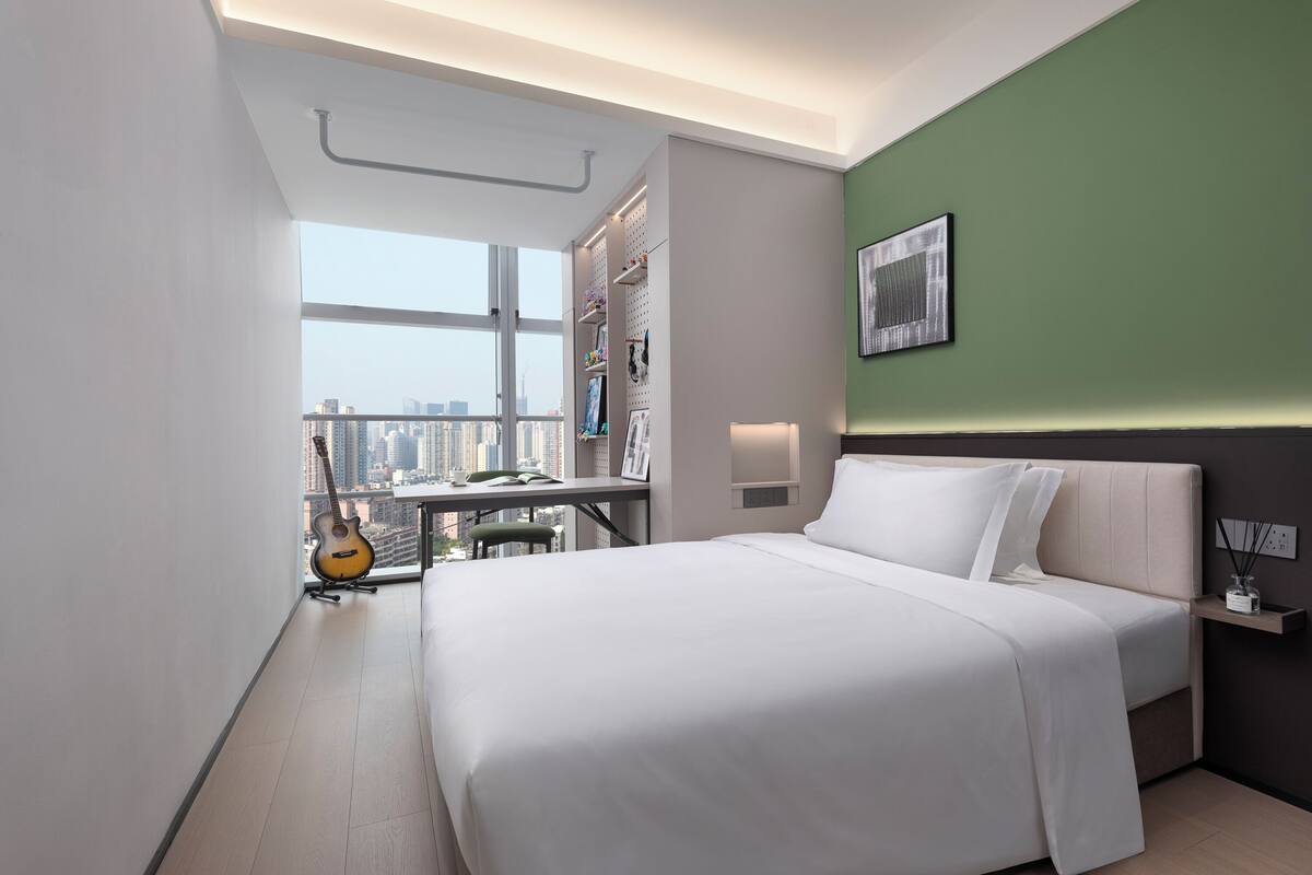 A modern hotel room with a large bed, a desk with a guitar, a city view, and a green accent wall.