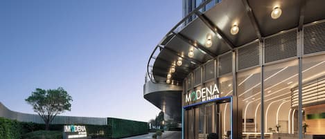 Una entrada de hotel moderna con fachada curva, ventanas amplias y un interior bien iluminado visible a través de las puertas de vidrio.