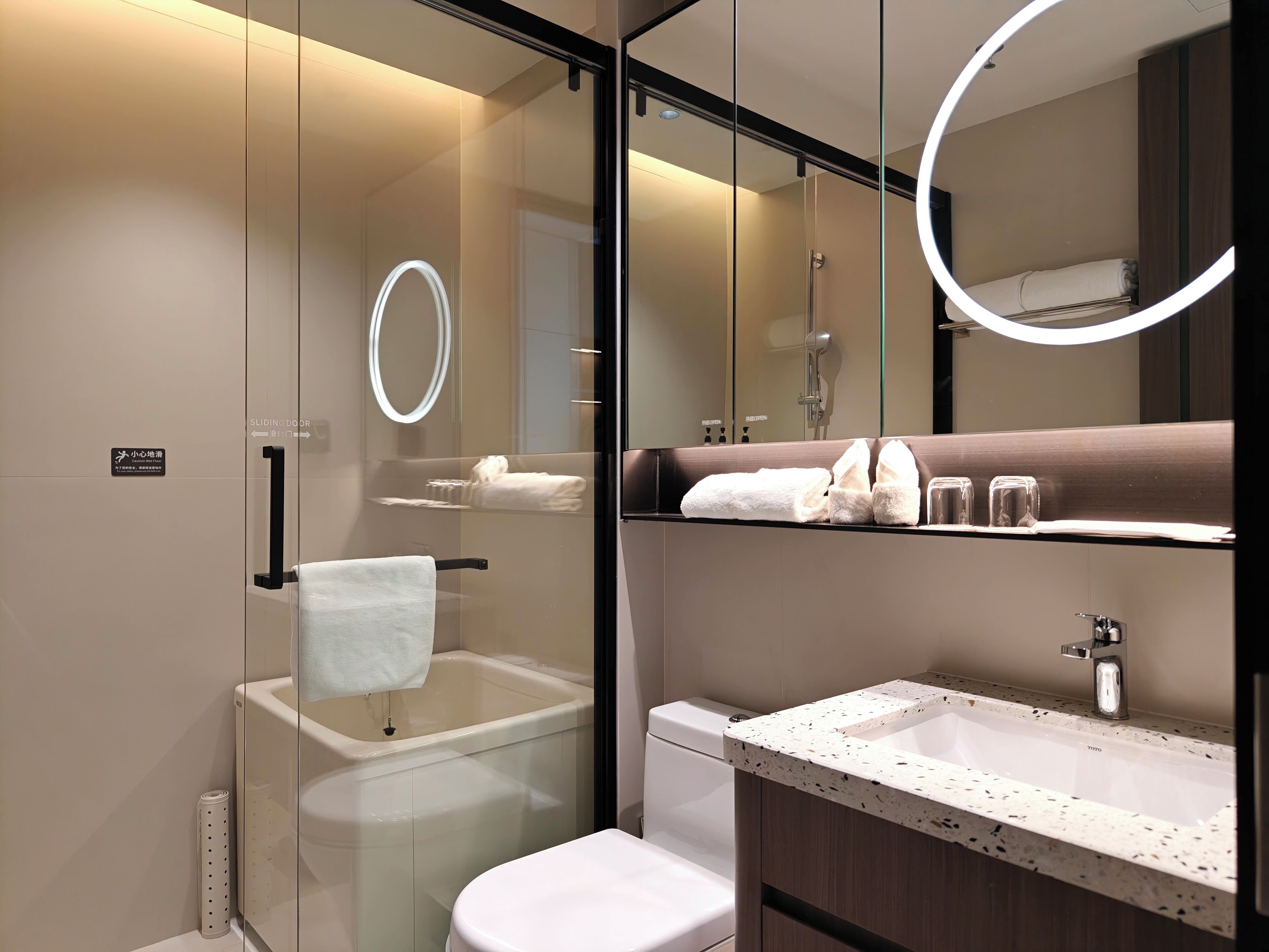 Une salle de bain moderne dotée d’une douche à parois de verre, de toilettes blanches, d’un lavabo avec un plan de travail à motifs et d’un miroir avec un éclairage circulaire.