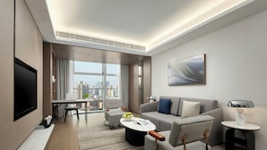 A modern hotel room with a large window, a flat-screen TV, a sofa, a coffee table, and a small side table with a lamp. - Modena By Freser Shenzhen (Shenzhen)