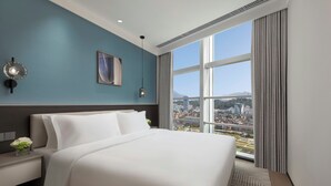 A modern hotel room with a large bed, a nightstand, a wall-mounted lamp, a framed picture, and a view of a cityscape through large windows. - Modena By Freser Shenzhen (Shenzhen)