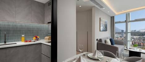 A modern hotel room with a dining area, a kitchen, and a large window offering a city view.