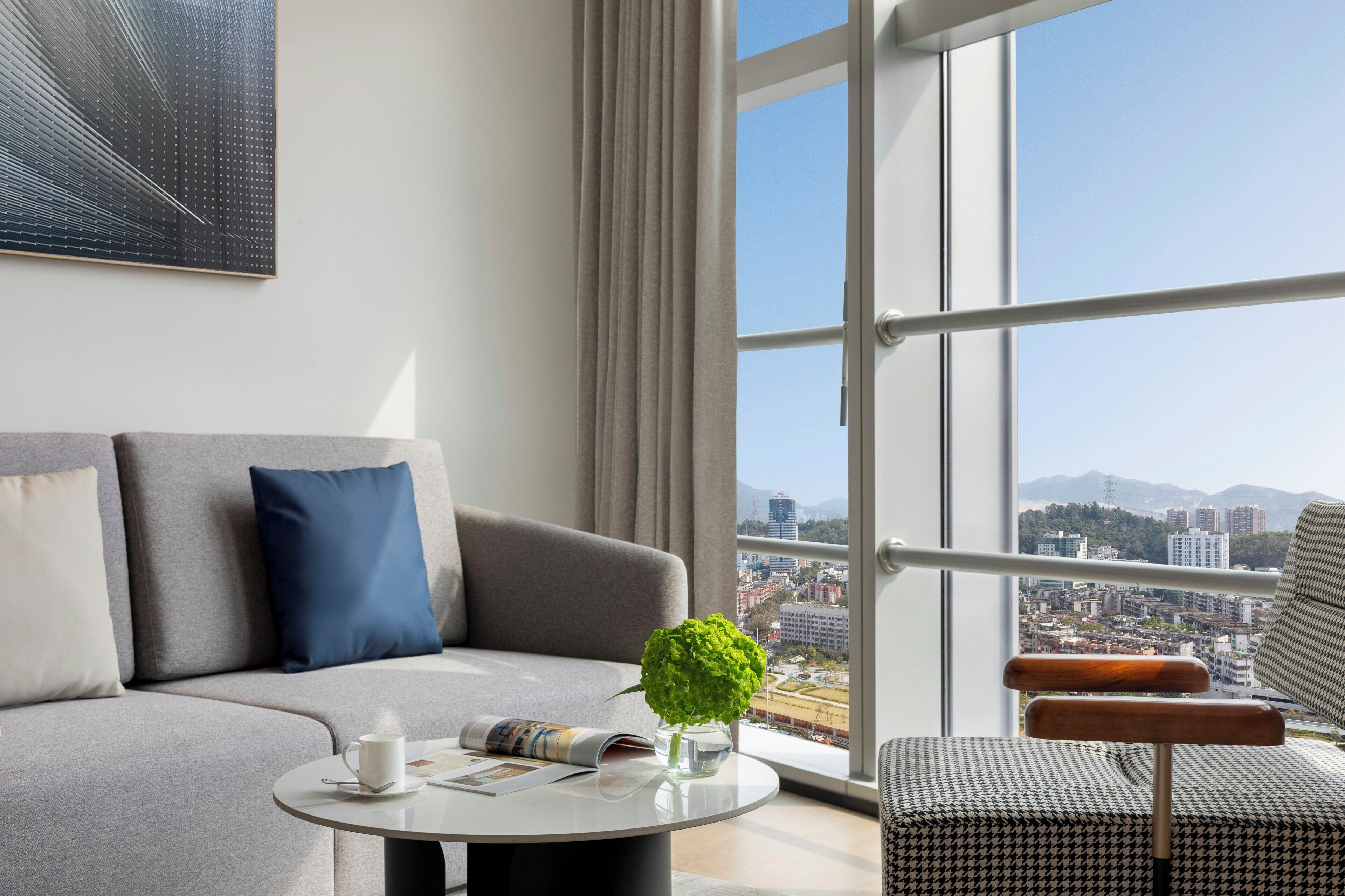 A modern living room with a grey sofa, a round coffee table, and a view of a cityscape through large windows.