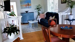 Un salon avec un plancher en bois, un escalier blanc, une table à manger avec des chaises, un canapé bleu et un téléviseur posé sur un meuble.