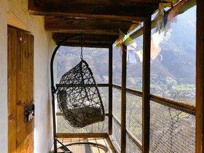 Une pièce avec un fauteuil suspendu, une porte en bois et une vue sur un paysage montagneux, visible à travers une fenêtre recouverte d’un treillis métallique.