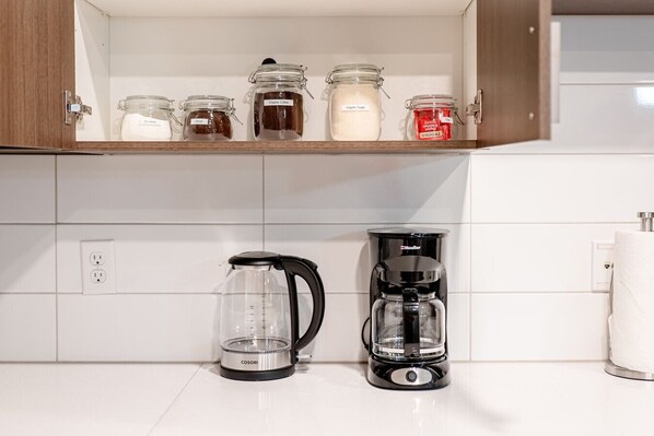 A kitchen with a coffee maker, a scale, and jars of coffee and sugar on a shelf.
