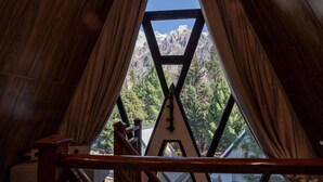 A view through a triangular window with a wooden frame, leading to a mountainous landscape with trees and a clear sky.